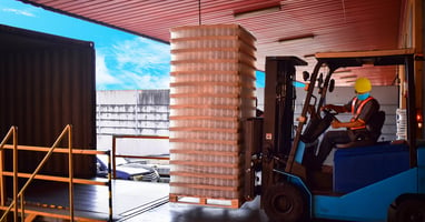 A worker wears a hard hat and operates a forklift. He moves products on pallets into a container on a warehouse dock.