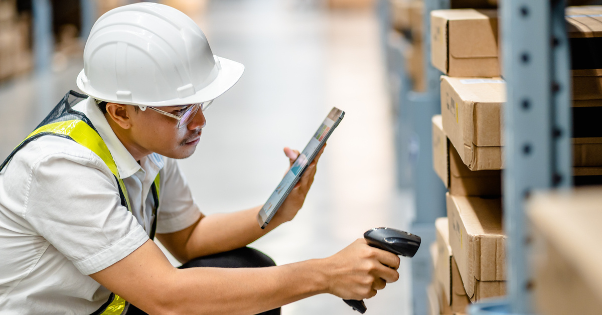 A warehouse worker wears a vest and a hard hat. He kneels down, scans a barcode on a box, and reads information on a tablet.