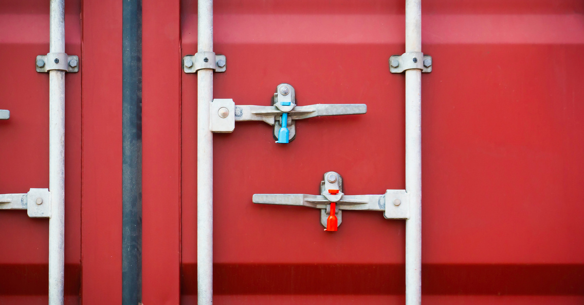 A cherry red cargo container is locked shut with metal posts. It's reinforced with two security seals in the mechanism.