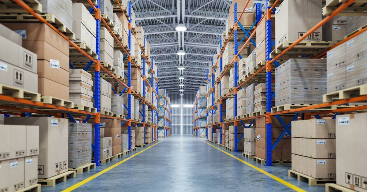 An aisle of a warehouse with four shelves filled with stacked cardboard boxes. The room is bright and the walkway is empty.