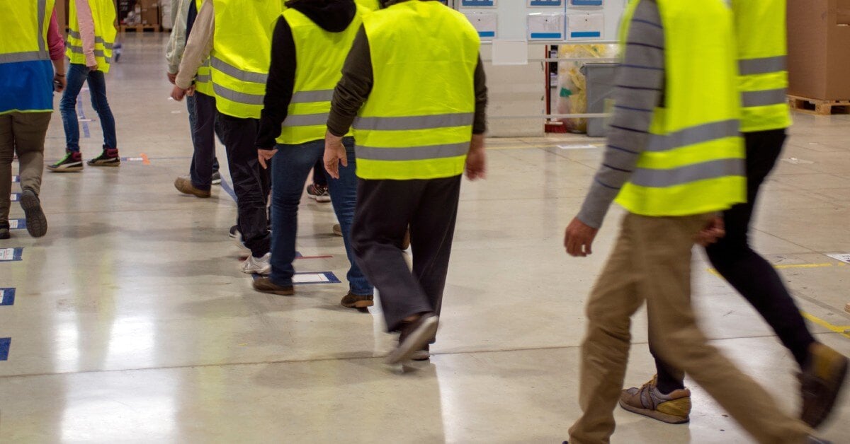 Eight men wearing pants and yellow reflective vests walk through a warehouse with sticker squares on the concrete floor.