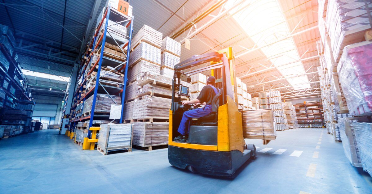 A forklift driver moves a pallet through the wide aisles of a warehouse. The shelves are stocked with goods.