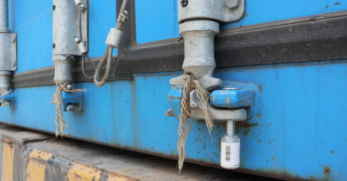 A warehouse loading dock floor has worn yellow stripes. A bolt seal with a barcode secures a blue shipping container.