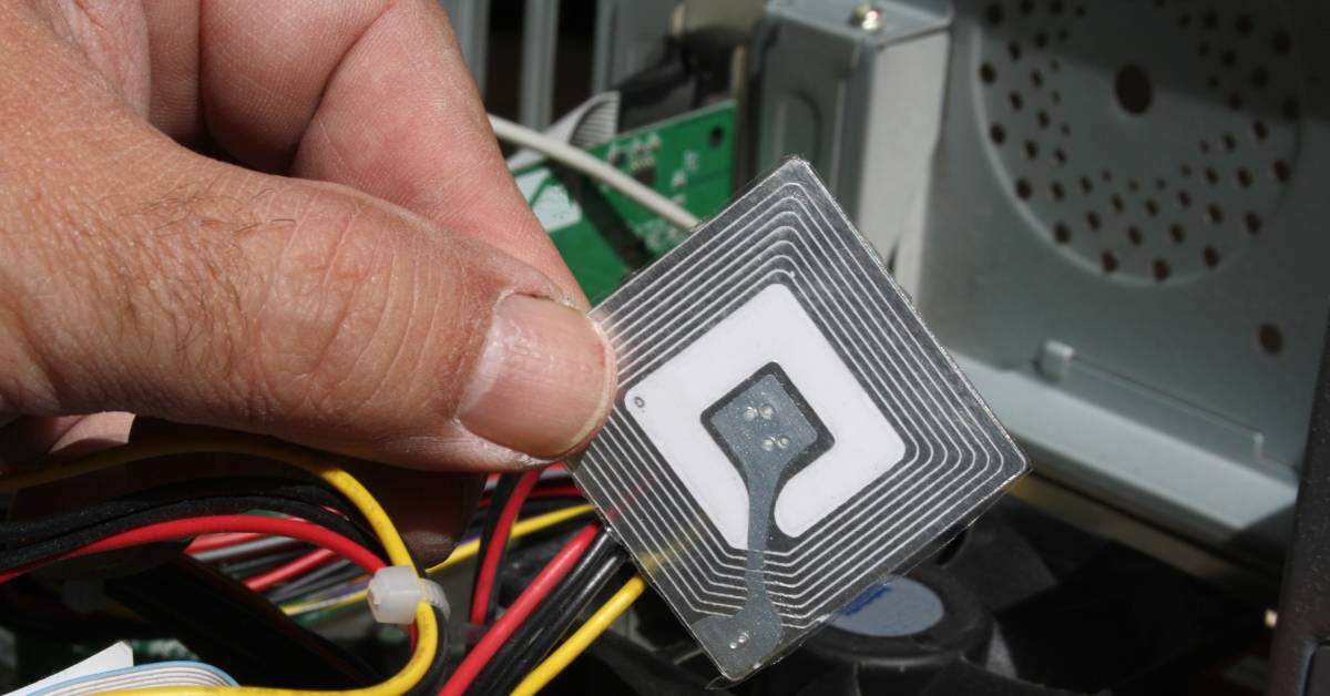 A hand is holding a small, silver, square RFID chip over a messy pile of yellow, red, and black wires.