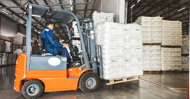 A man wearing a blue jumpsuit operates a forklift in a warehouse unloading a large stock of goods in white packaging.