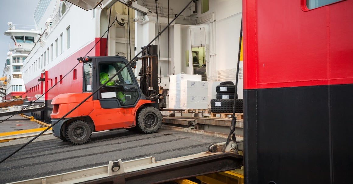 A forklift drives up a ramp and collects a pallet of white boxes on a loading dock outside of a warehouse.