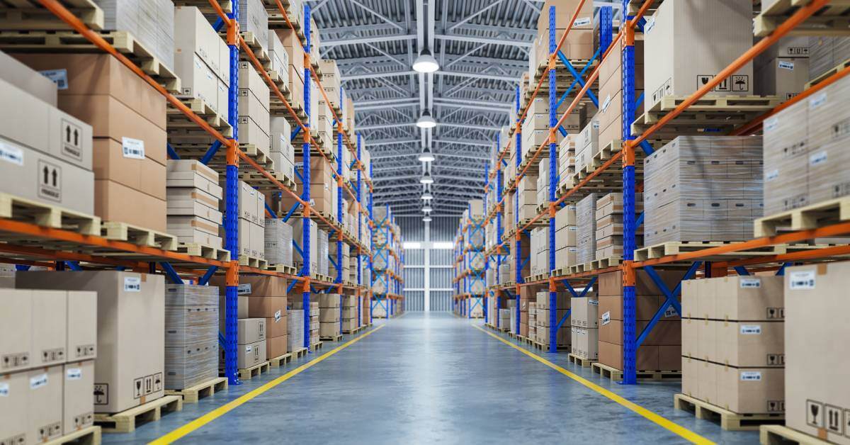 A large warehouse aisle with orange and blue shelving racks. The racks hold boxes with pallets underneath on both sides.