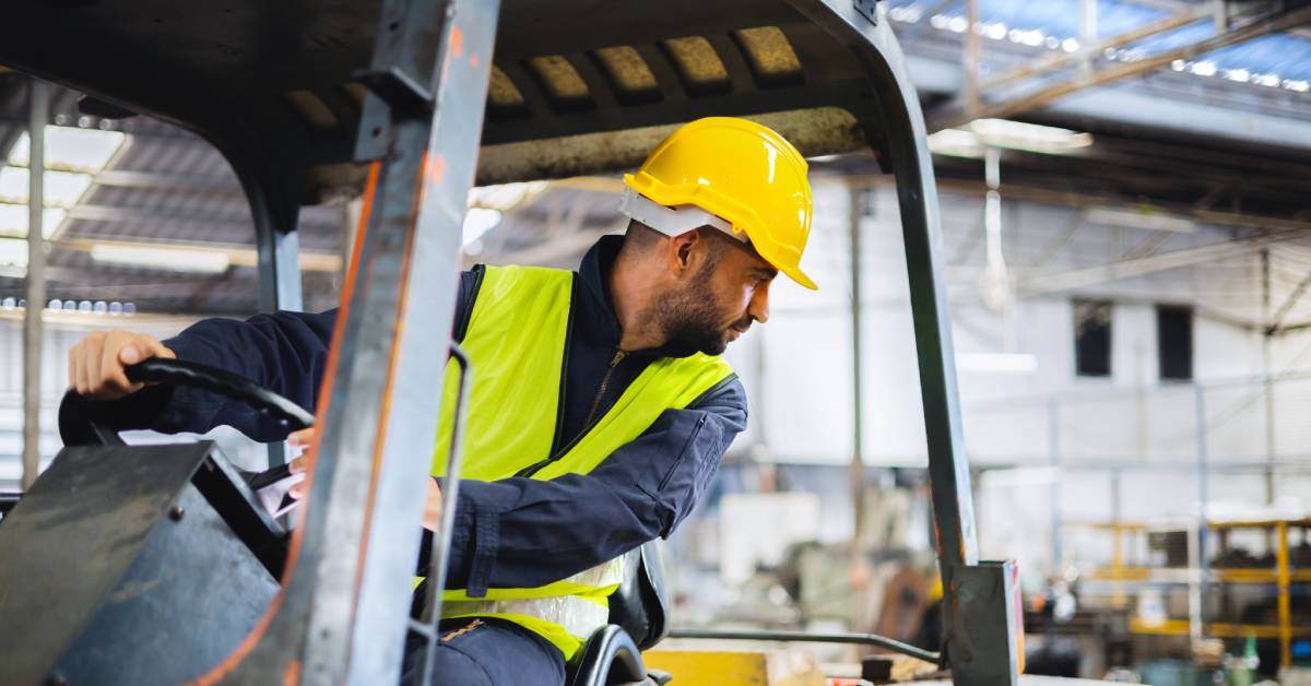 A worker wearing a hard hat and a bright vest operates a forklift and looks backward in a warehouse.