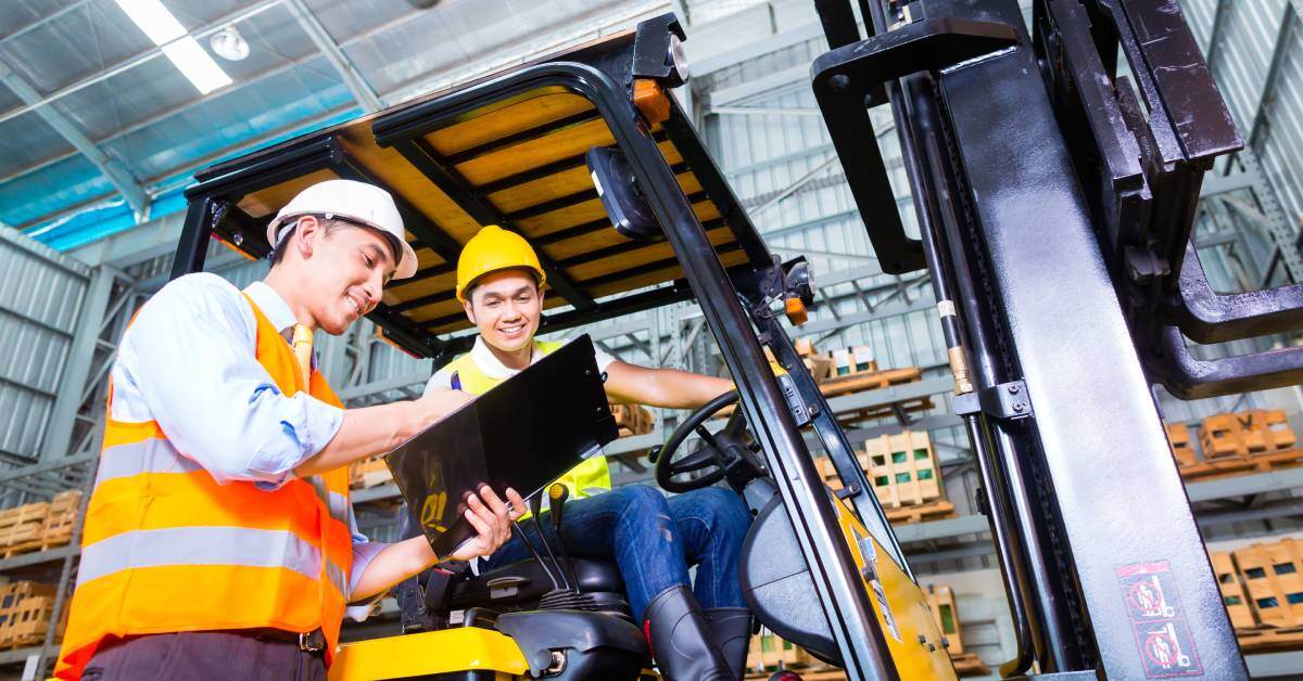 One man wearing a hard hat and vest holding a clipboard speaks to another who's operating a forklift.