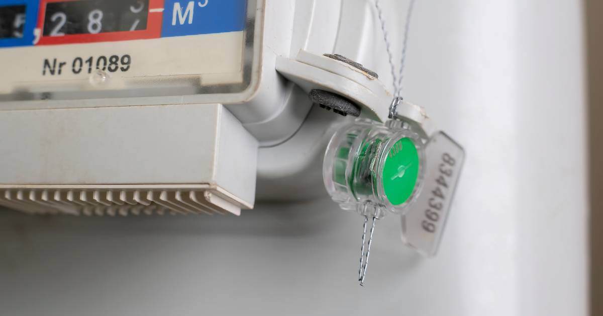 A gas meter located on a white wall, with a wire and green plastic seal that protect the meter from tampering.