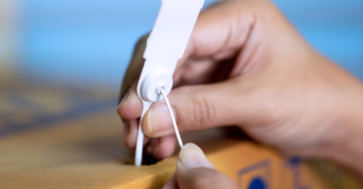 Someone's hand is placing a white plastic tamper-evident security seal on the handle of a corrugated box.