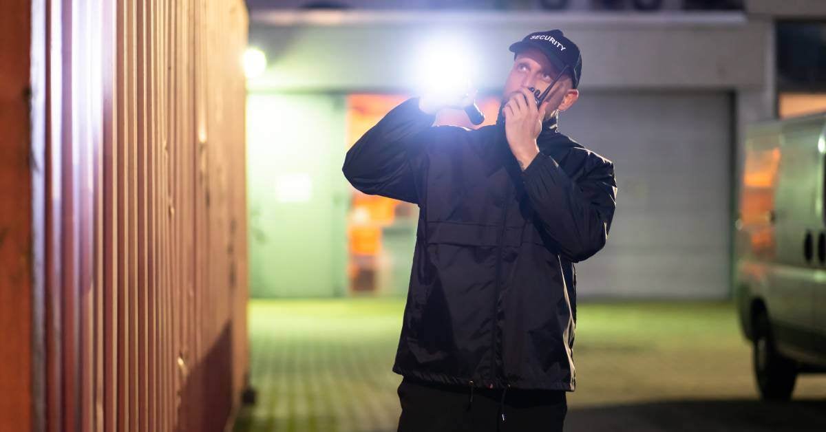 A security guard in a black jacket and hat points a flashlight at a shipping container. He is speaking on a two-way radio.