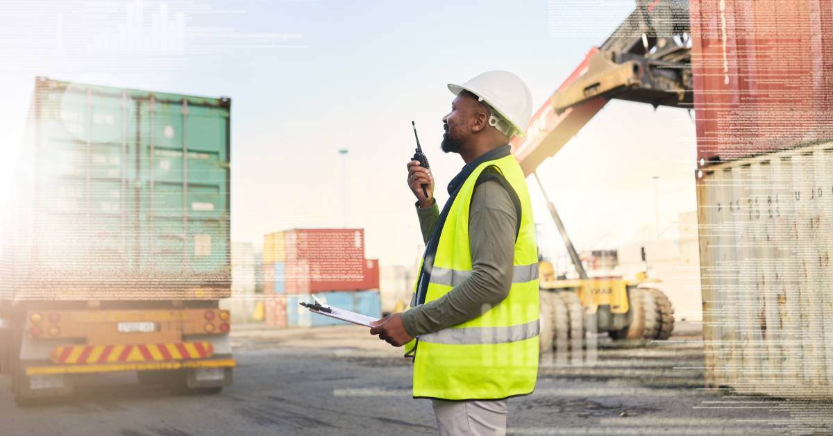 A man wearing a hard hat and reflective vest speaks into a walkie-talkie at a cargo warehouse with stacked shipping containers.