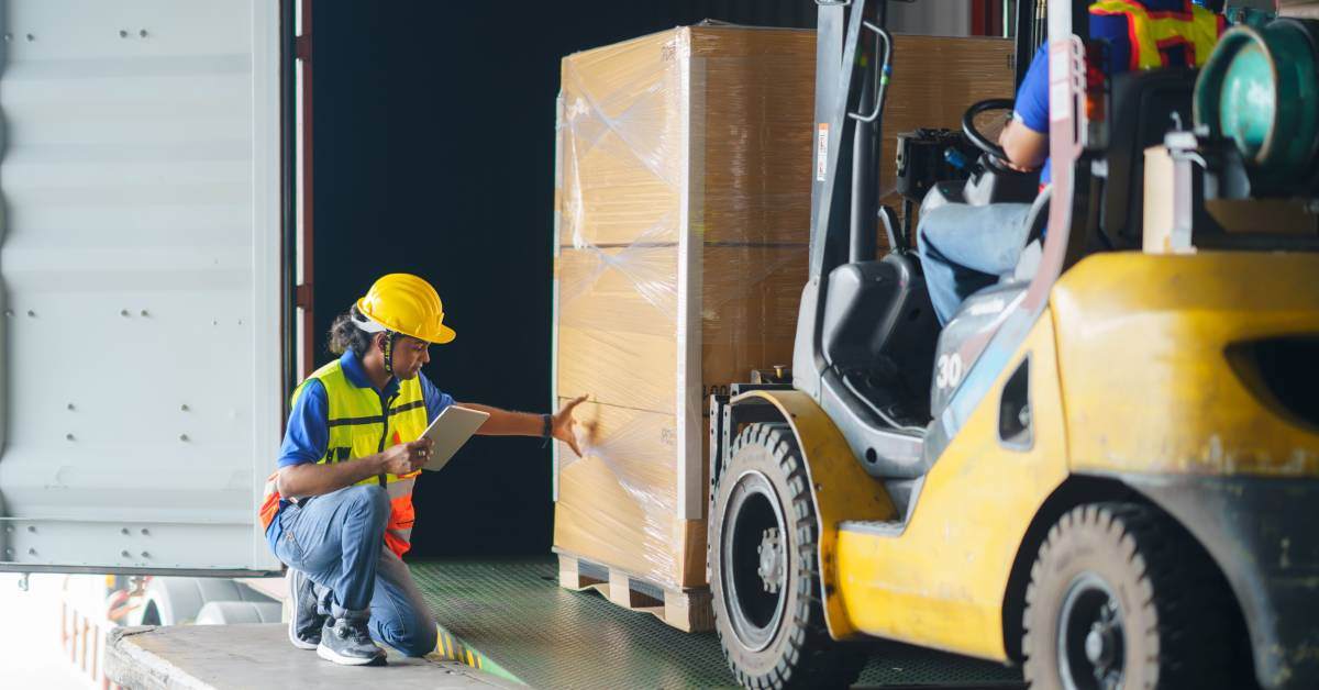 A warehouse worker holding a notepad, wearing a hard helmet and a bright vest, inspects boxes on a forklift.