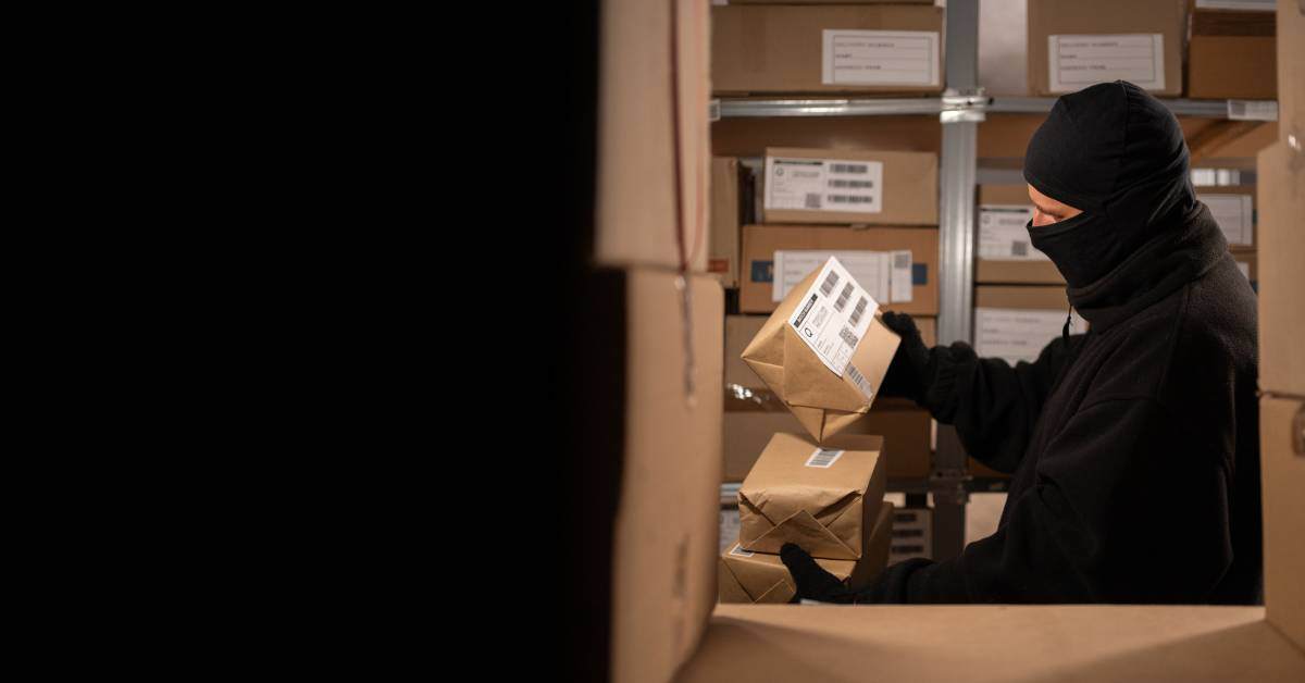 A thief dressed in all black and a ski mask is between stacks of packages. They are holding and looking at multiple packages.